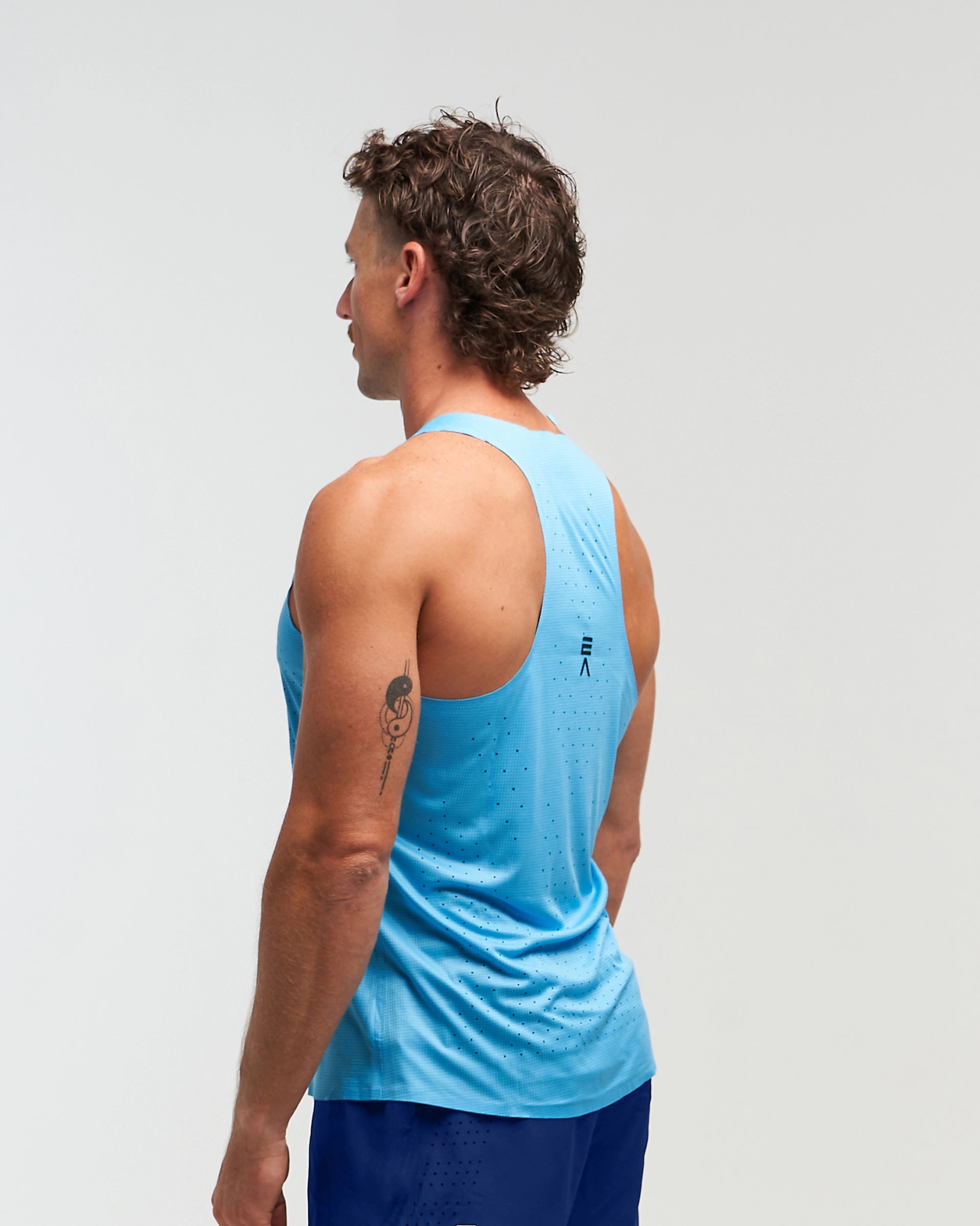 Side pose with back visible of man wearing ocean blue EVOActive Men’s Aero Singlet with logo visible against a bright background