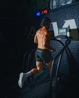 Man wearing midnight black EVOActive Unisex Aero Run Short running on a treadmill in a dark gym setting with logo visible on the shorts