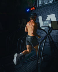 Man wearing midnight black EVOActive Unisex Aero Run Short running on a treadmill in a dark gym setting with logo visible on the shorts