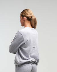 Back view of woman wearing clouded grey EVOActive Unisex Classic LS Crew Neck Tee with logo visible against a bright background