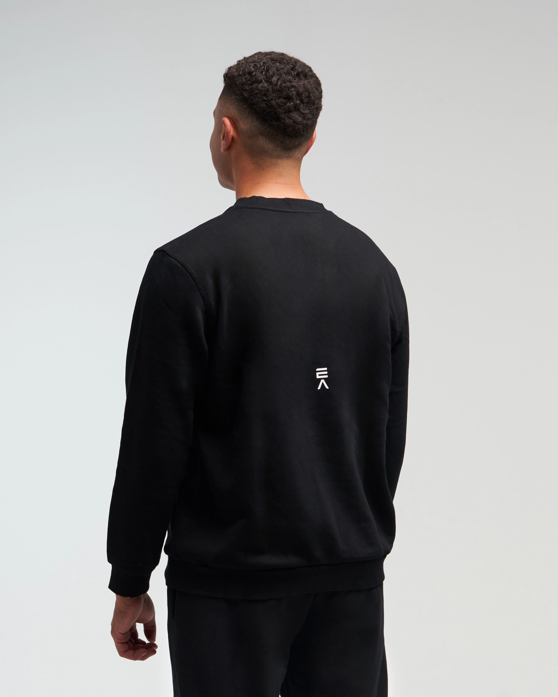 Back view of man wearing midnight black EVOActive Unisex Classic LS Crew Neck Tee with logo visible on the garment against a bright background
