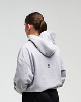 Back view of woman in a slight side pose wearing clouded grey EVOActive Unisex Classic Pullover Hoody with logo visible on the garment against a bright background