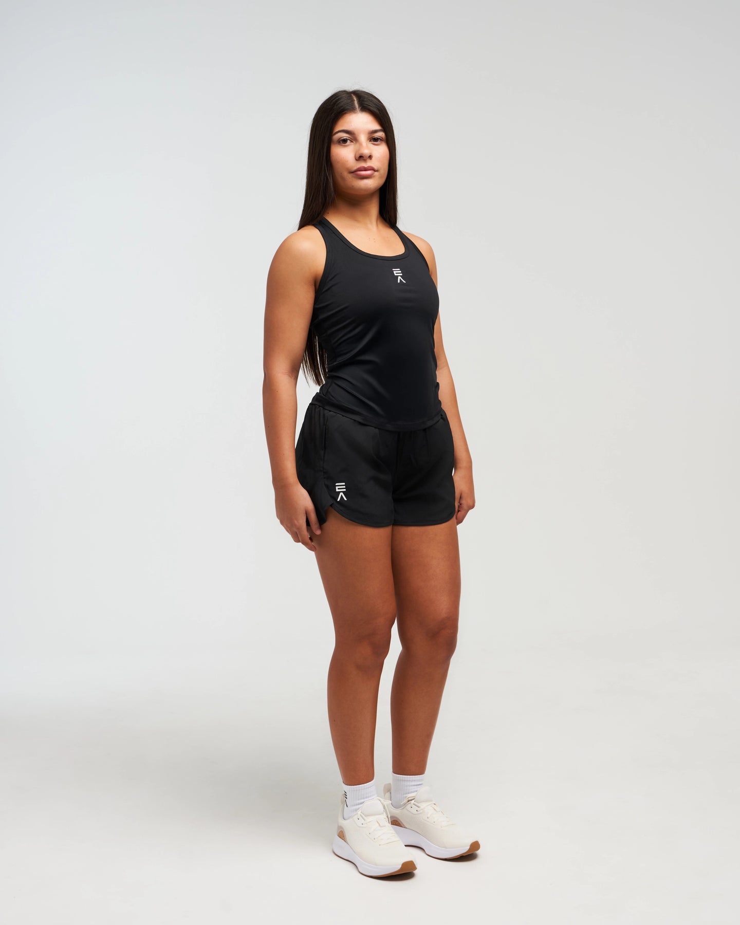 Full body front view of woman wearing midnight black EVOActive Women’s CoreRacer Short with logo visible against a bright background