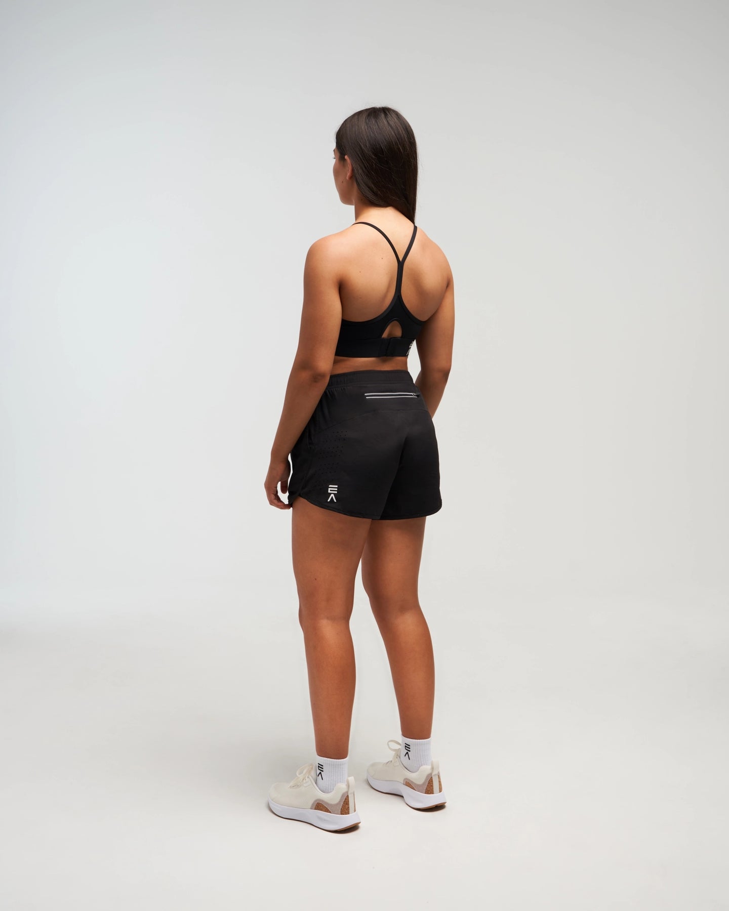 Full body back view of woman wearing midnight black EVOActive Women’s Flex Sports Bra in a slight side pose with logo visible against a bright background