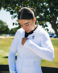 Woman wearing Evo Ice white EVOActive Women’s Horizon Jacket zipping it up while standing against green tree background with logo visible