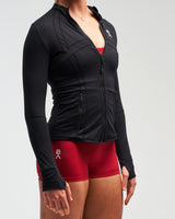 Mid-body view of woman wearing midnight black EVOActive Women’s Horizon Jacket with logo visible against bright background