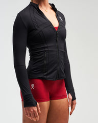 Mid-body view of woman wearing midnight black EVOActive Women’s Horizon Jacket with logo visible against bright background