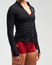 Mid-body view of woman wearing midnight black EVOActive Women’s Horizon Jacket with logo visible against bright background
