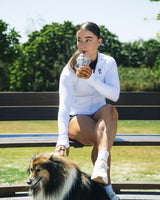 Woman sitting on a bench wearing Evo Ice white EVOActive Women’s Horizon Jacket, sipping a beverage with dog nearby and logo visible against trees and open field background