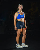 Full body front view of woman wearing black EVOActive Women’s Motion Pocket Short in slight side pose with blue sports bra against dark background

